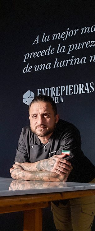 Hombre con tatuajes, apoyado en una mesa, sonríe frente a un fondo oscuro. con marca HARINAS ENTREPIEDRAS